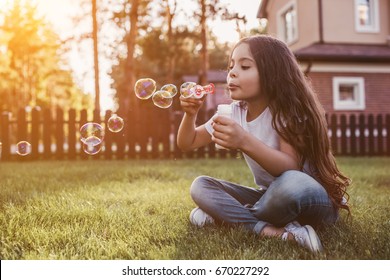 Pretty little girl is sitting on the green grass on the backyard and blowing soap bubbles. Having fun on the sunset.