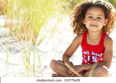 Pretty little girl sitting in the sand while playing with the tall grass near the ocean