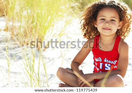 Pretty little girl sitting in the sand while playing with the tall grass near the ocean