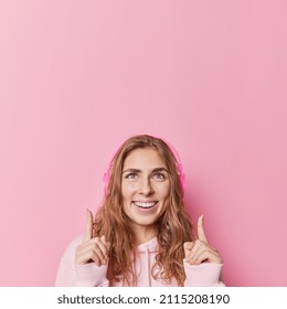 Pretty Long Haired Woman Smiles Gladfully Dressed In Casual Sweatshirt Listens Favorite Melody Points Index Fingers Overhead Isolated Over Pink Background Suggests To Click On Link. Look At This