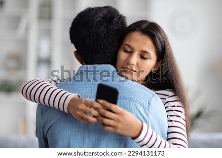 Pretty long-haired young eastern woman chatting with guys on dating app via smartphone while hugging her boyfriend, home interior, copy space. Cheating, deception, dishonesty in relationships