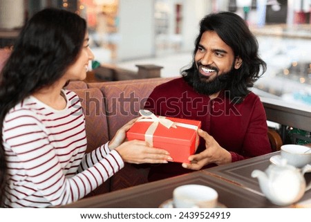 Pretty long-haired young indian woman presenting wrapped gift box to her excited boyfriend at cafe, making special surprise. Loving couple celebrating birthday or anniversary at coffee shop