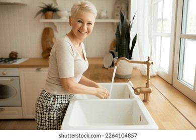 Pretty mature woman with short blonde hair washing something in white kitchen sink, preparing meal or cleaning place after dinner, looking at camera smiling happily. Domestic life. Daily routine - Powered by Shutterstock