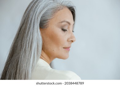 Pretty middle aged Asian woman with loose straight grey hair poses on light background in studio closeup side view. Mature beauty lifestyle