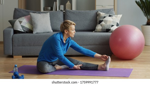 Pretty middle aged woman in sporty style with short blond hair sitting on the yoga mat and stretching her legs and back in the living room. Indoor - Powered by Shutterstock