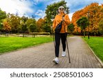 Pretty middle-aged blonde woman dressed sporty practicing Nordic walking on background of beautiful autumnal trees in city park on autumn day. Front view. Autumnal training of Nordic walking.