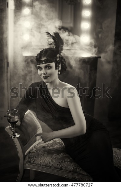 Pretty model.\
Flapper girl with wineglass. Old photo in a retro style. Smoky\
eyes, hairstyle cold wave. Creative colors. Hair ornament with\
ostrich feathers, lace\
bandage.
