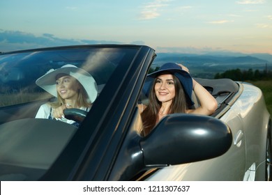 Pretty Models In Dark Blue And White Brim Broad Hats Sitting On Front Seats Of Silver Cabriolet. Adorable Girls Driving Car, Looking Away And Smiling. Concept Of Luxurious Automobile And Journey.