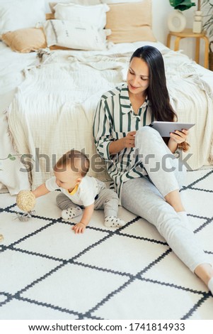 A pretty mom with a computer tablet sits on the floor and spends time in pleasure playing with a toddler baby. Remote work and study online. Social distance and business in the coronovirus pandemic.
