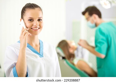 Pretty Nurse Is Speaking By Phone And Looking At Camera With Smile. Dentist And Patient On Background.