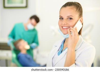 Pretty Nurse Is Speaking By Phone And Looking At Camera With Smile. Dentist And Patient On Background.