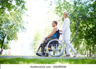 Pretty Nurse Walking With Male Patient In A Wheelchair In Park