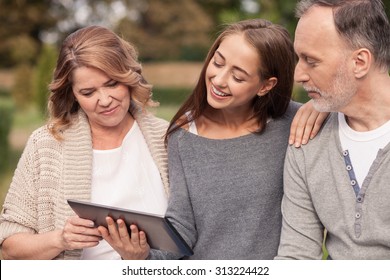 Pretty old married couple and their young daughter are using a tablet. They are looking at it with interest and smiling. They are sitting in park and embracing with love స్టాక్ ఫోటో