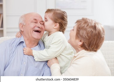Pretty old men and women are spending time with their granddaughter. The girl is kissing her grandfather with joy. They are sitting on sofa and laughing