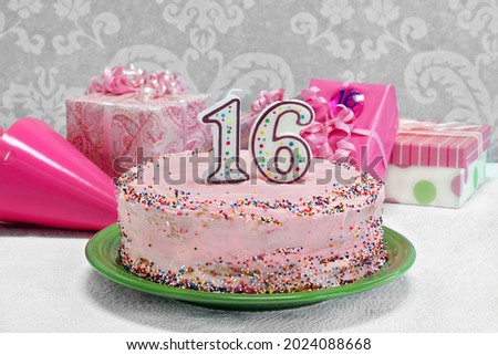 A pretty pink, sprinkled birthday cake with the numbers 16 on top.  Gifts and party hats by cake.