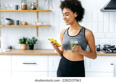 Pretty, positive African American curly girl stands at home in the kitchen, holds a glass of fresh organic green smoothie in her hand, uses her smartphone, messaging on social networks, smiles - Powered by Shutterstock