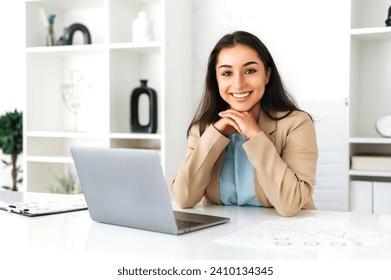 Pretty, positive successful brunette indian or arabian business woman in an elegant suit, ceo, company employee, sitting at a work desk in the modern office, looking at camera and smiling friendly - Powered by Shutterstock