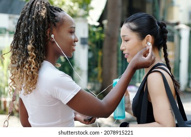 Pretty Positive Young Woman Sharing Earbuds With Friend So They Can Listen To Tranding Music Track