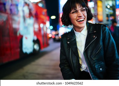Pretty positive young woman with short haircut laughing in urban setting with night lights enjoying leisure time in New York.Happy hipster in stylish wear and eyewear having fun on megapolis street - Powered by Shutterstock