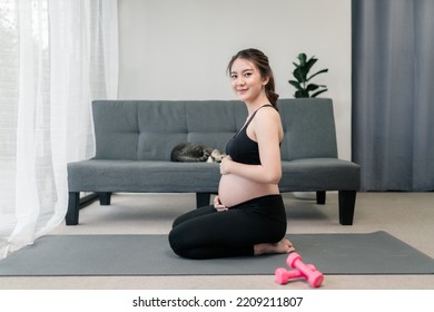 Pretty Pregnant Asian Woman Is Sitting With A Smile On Her Face While Looking At The Camera And Her Hands Rubbing Her Pregnant Tummy. Concept Healthy Pregnancy.