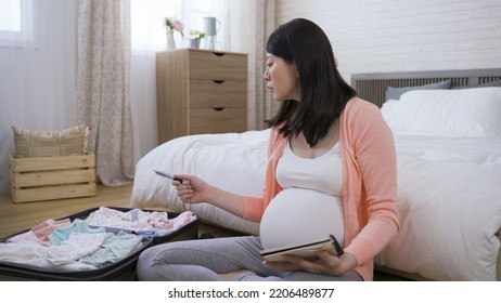 Pretty Pregnant Lady Sitting Against The Bed On Floor At Home Is Counting The Baby Items With A Pen In Hand And Looking At Her Supply Checklist.