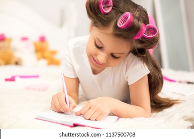 Pretty Preschooler Writing In Diary On White Bed. Sweet Girl With Hair Rollers Tilting Her Head To Side While Writing Down Some Notes In Her Journal.