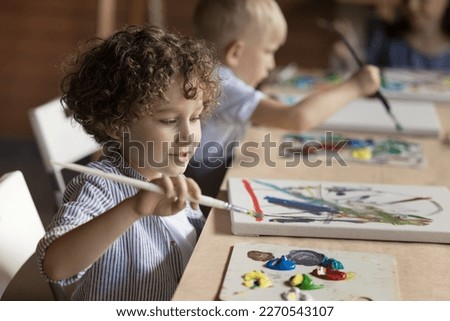 Pretty pupil kid boy painting on class, studying art in artistic school for children, drawing abstract picture on canvas at table, using paintbrush, palette with oil, acrylic paints