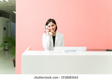 Pretty receptionist in a white robe talking on the phone at reception desk of the doctor's office. Female worker smiling and taking a medical appointment of a patient - Powered by Shutterstock