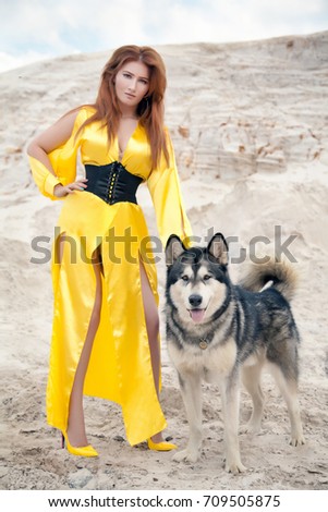 pretty red hair woman wearing long evening yellow dress and her lovely big happy dog malamute in the desert quarry white sand