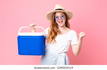 mujer de la cabeza roja guapa sintiéndose impactada, riendo y celebrando el éxito y sosteniendo un refrigerador portátil de picnic