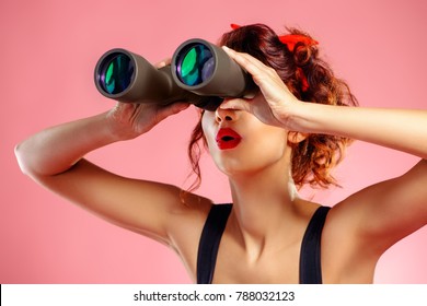 Pretty red-haired girl looks through binoculars at something over pink background. Pin-up style. 