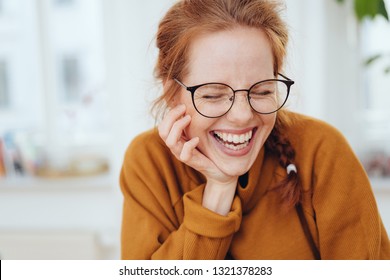 Linda garota ruiva com rabo de cavalo, usando óculos e moletom laranja, rindo com os olhos fechados. Retrato frontal de close-up dentro de casa com espaço de cópia