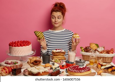 Pretty Redhead Woman Chooses Between Croissant And Cake, Sits At Table Overloaded With Desserts, Dressed In Striped Jumper, Isolated Over Pink Wall. Unhealthy Eating, Harmful Food And Sugar Obsession