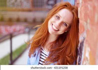 Pretty redhead woman with a lovely friendly smile leaning against a brick wall in an urban environment beaming at the camera, close up head and shoulders