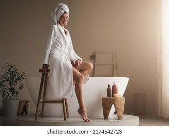 Pretty relaxed female having rest after taking bath with aroma oil, sitting next to ceramic bathtub on wooden stool in bathrobe and towel, looking aside smiling. Body care concept - Powered by Shutterstock