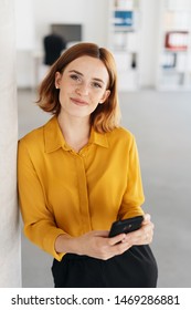 Pretty relaxed young woman with friendly smile leaning against an interior white wall holding a mobile phone