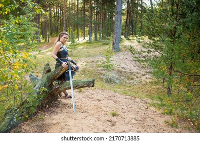 Pretty Robber Girl Wearing Black Leather Shoulder Guard, Skirt, Lorica And  Bracer With Steel Estramacon Is Resting On The Fallen Tree In A Summer Forest