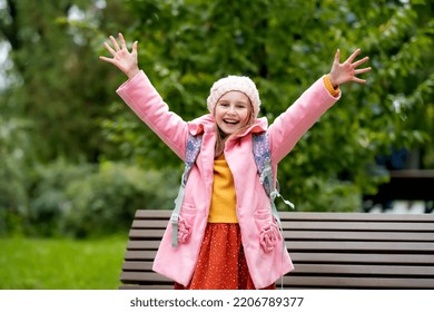 Pretty School Girl Kid With Backpack Looking At Camera, Holding Hands Up And Smiling At Autumn Park. Beautiful Female Child Portrait