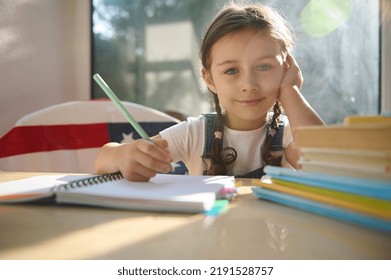 Pretty Schoolgirl, Adorable Caucasian Child Holding A Pencil, Learning To Write, Sitting At Desk With Stacked Textbooks, Cutely Smiles Looking At Camera. American Flag On The Chair On The Background