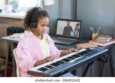 Pretty schoolgirl in headphones playing piano keyboard while listening to advice of her teacher during online lesson