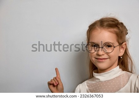 Pretty schoolgirl in a white blouse and round glasses points her finger at the school blackboard. Little girl in round glasses with a black frame stands against a white wall, advertising something