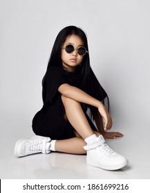 Pretty Self-confident Early Adult Serious Brutal Asian Kid Girl In Stylish Black Dress, White Sneakers And Sunglasses Sits On Floor Cool Posing. Trendy Children Fashion Concept