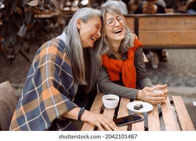 Pretty senior Asian woman puts head on laughing friend shoulder resting together at table in street cafe on nice autumn day - Shutterstock ID 2042101487