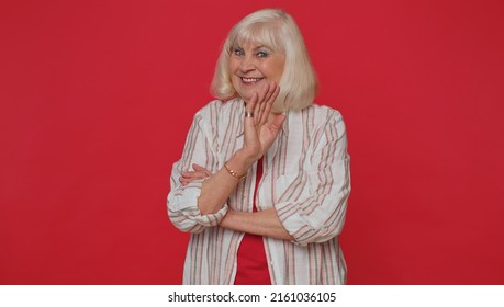 Pretty Senior Old Woman In Shirt Smiling Friendly At Camera And Waving Hands Gesturing Hello Or Goodbye, Welcoming With Hospitable Expression. Happy Grandmother Indoors Isolated On Red Wall Background