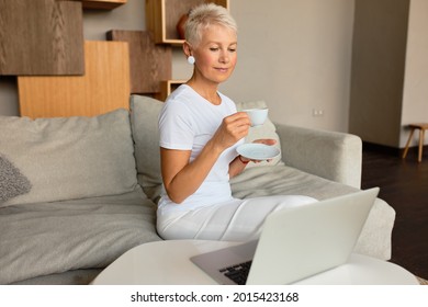 Pretty senior woman enjoy tea from porcelain tableware. Lady watch interesting movie on laptop resting on couch at home. Leisure, pastime, cinema, time alone, lazy sunday concept - Powered by Shutterstock