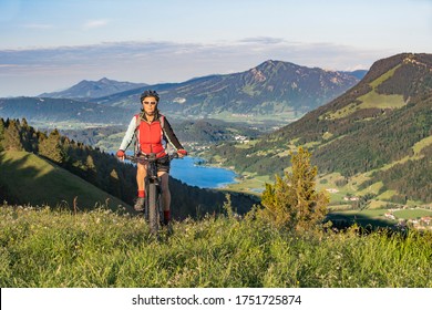 pretty senior woman riding her electric mountain bike in warm dawn sunlight heights of Salmas hight above Oberstaufen, with spectacular view on lake Alpsee,Allgau Alps, Bavaria Germany 