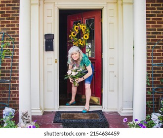 Pretty Senior Woman In Shorts And Tee-shirt At Door To Brick House With Pillars And Sunflower Wreath Picks Up Flower Delivery Left On Porch For Birthday Or Mothers Day