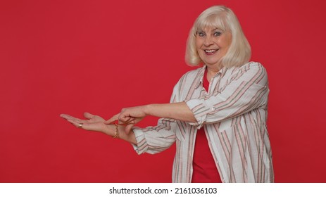 Pretty senior woman showing thumbs up and pointing empty place, advertising area for commercial text, copy space for goods promotion. Elderly grandmother. Studio shot indoors on red wall background
