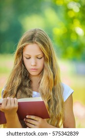 Pretty severe young girl reads red book, against green of summer park.