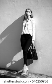 Pretty Sexy Cute Business Woman Or Girl With Long Hair In Pants Fashion Bag And Blouse On High Hells Posing Outdoor On Wall Background, Black And White